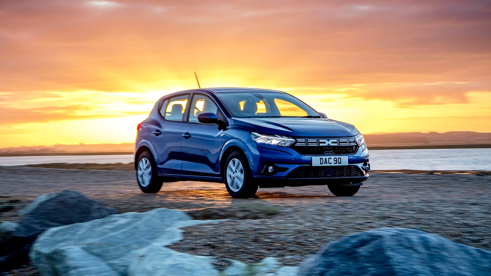 Dacia Sandero in blue, on a beach in front of a sunset